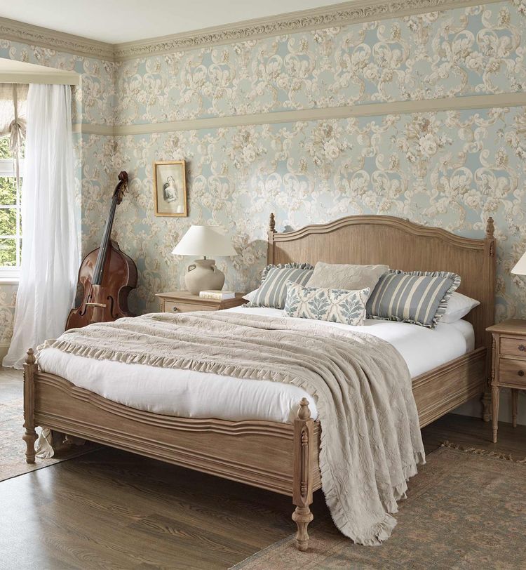 Elegant bedroom with a wooden bed, floral wallpaper, cello in the corner, and light streaming through a window with sheer curtains.