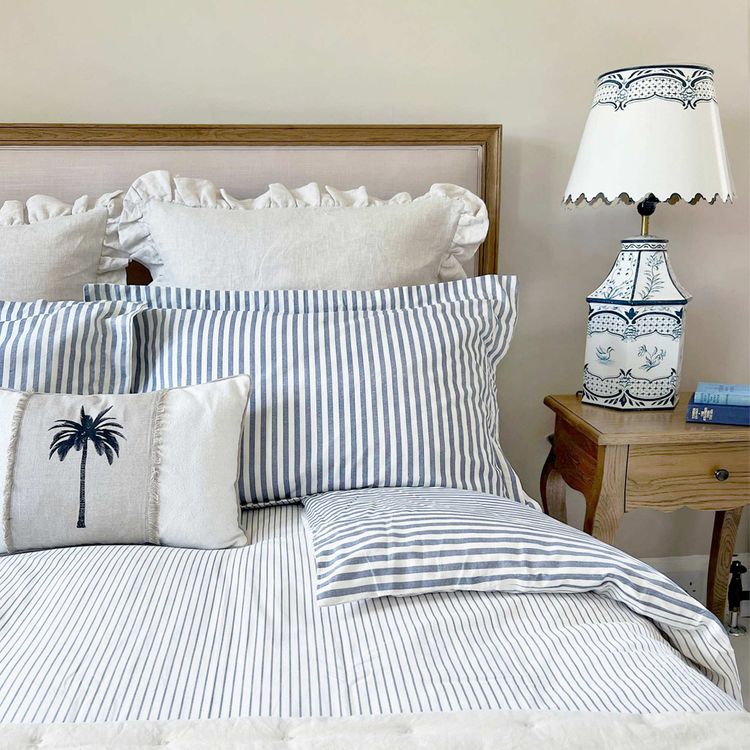 Cozy upholstered bed with blue striped sheets, palm tree cushion, ruffled pillows, and a wooden side table.