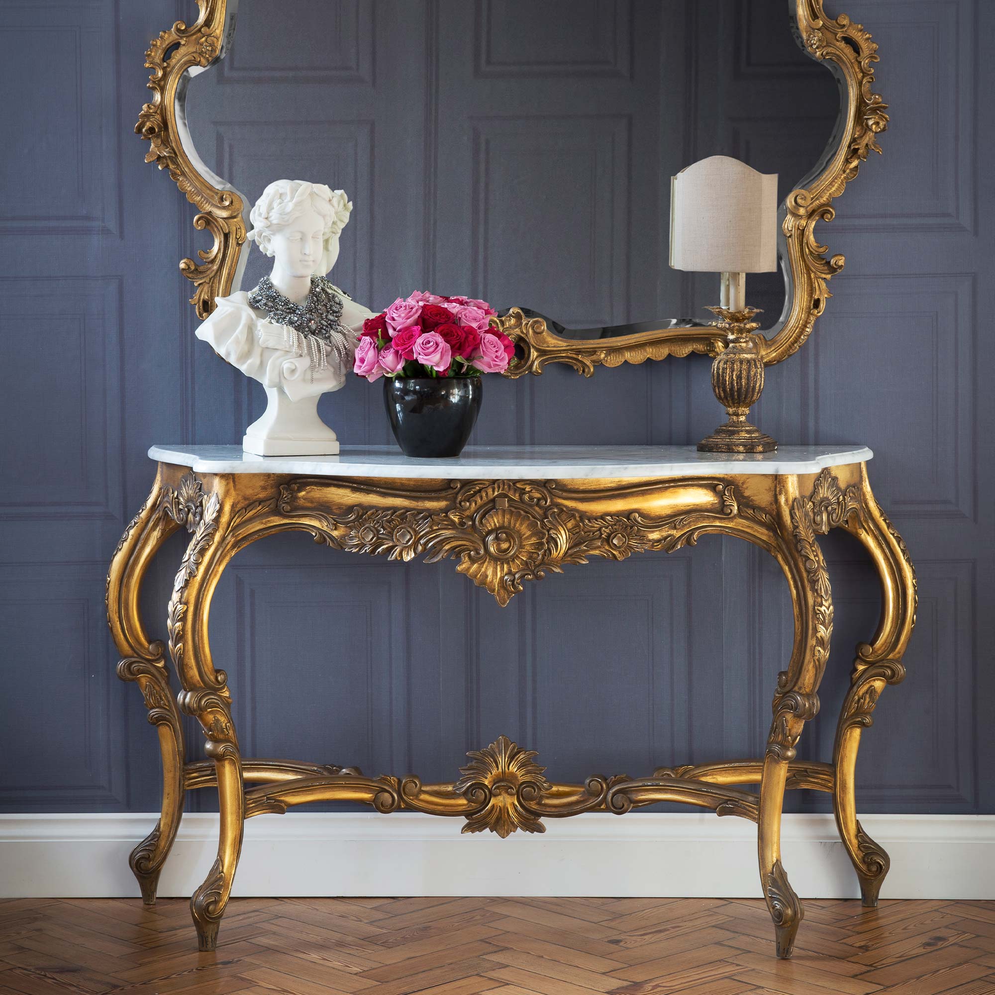 Ornate gold console table with a marble top, featuring a bust, vase of pink roses, and a lamp, set against a paneled gray wall and mirror.