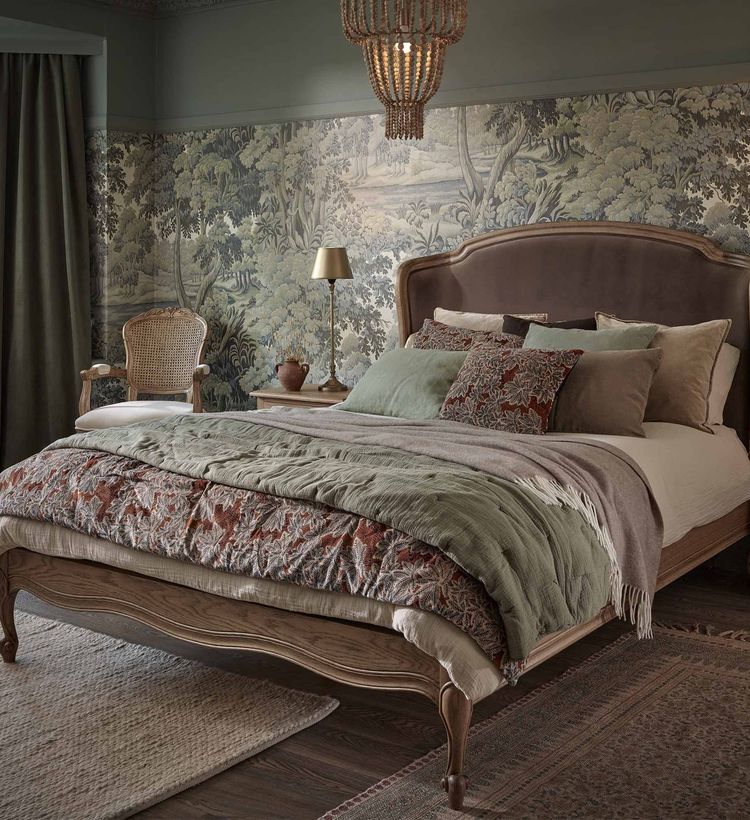 Elegant bedroom with a vintage bed, layered bedding, patterned wallpaper, a woven chair, and a chandelier, creating a cozy, classic ambiance.