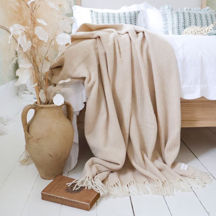 A cozy beige fringed cashmere blanket drapes over a bed next to a terracotta vase with dried flowers and a book titled "Wild Flowers" on a white floor.
