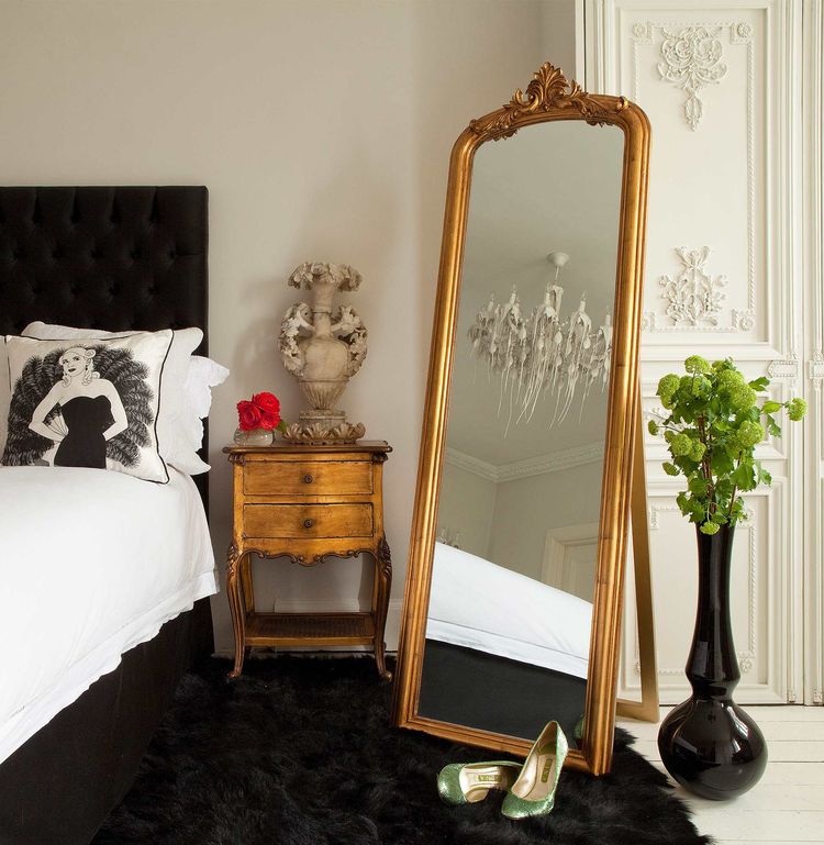 Elegant bedroom with a tall gold-framed mirror, vintage side table, decorative vase, black rug, and potted plant. Shoes placed near the mirror.