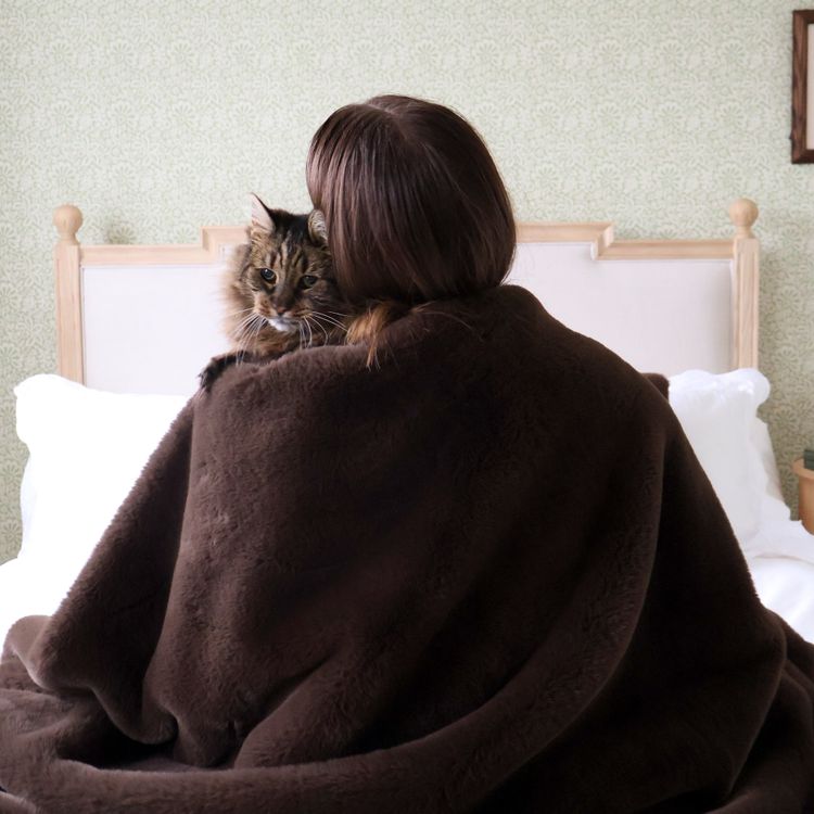 Person sitting on a bed, wrapped in a brown blanket, holding a cat on their shoulder, facing away.