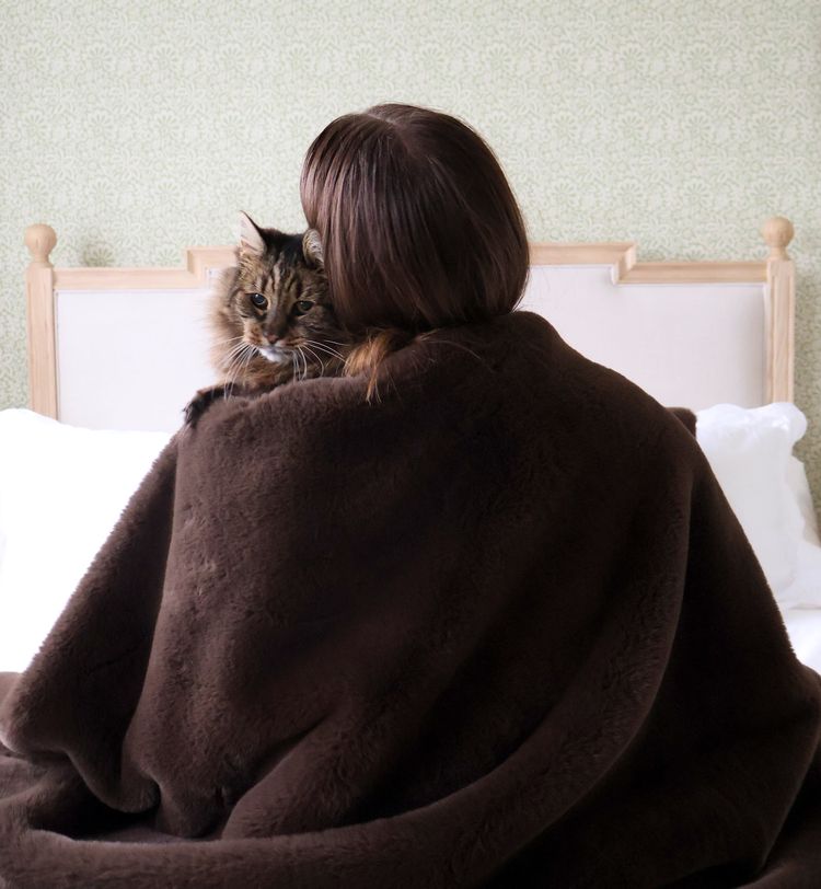 Person sitting on a bed, wrapped in a brown blanket, holding a cat on their shoulder, facing away.
