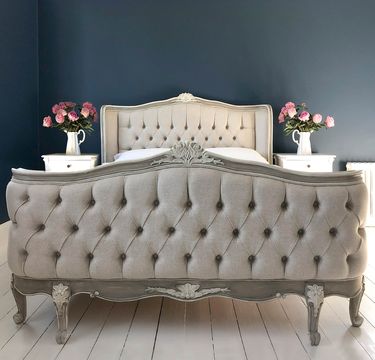 Elegant bedroom with a tufted, upholstered bed, flanked by white nightstands with pink flowers in vases, against a dark blue wall.