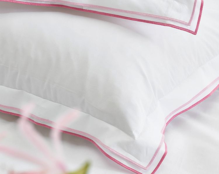 Two stacked white pillows with pink trim on a bed, featuring soft fabric and a blurred flower in the foreground.