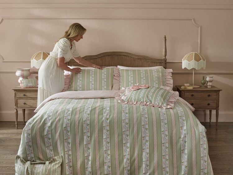 A woman arranges pillows on a bed with pink and green striped bedding in a softly lit bedroom with matching decor and wooden furniture.