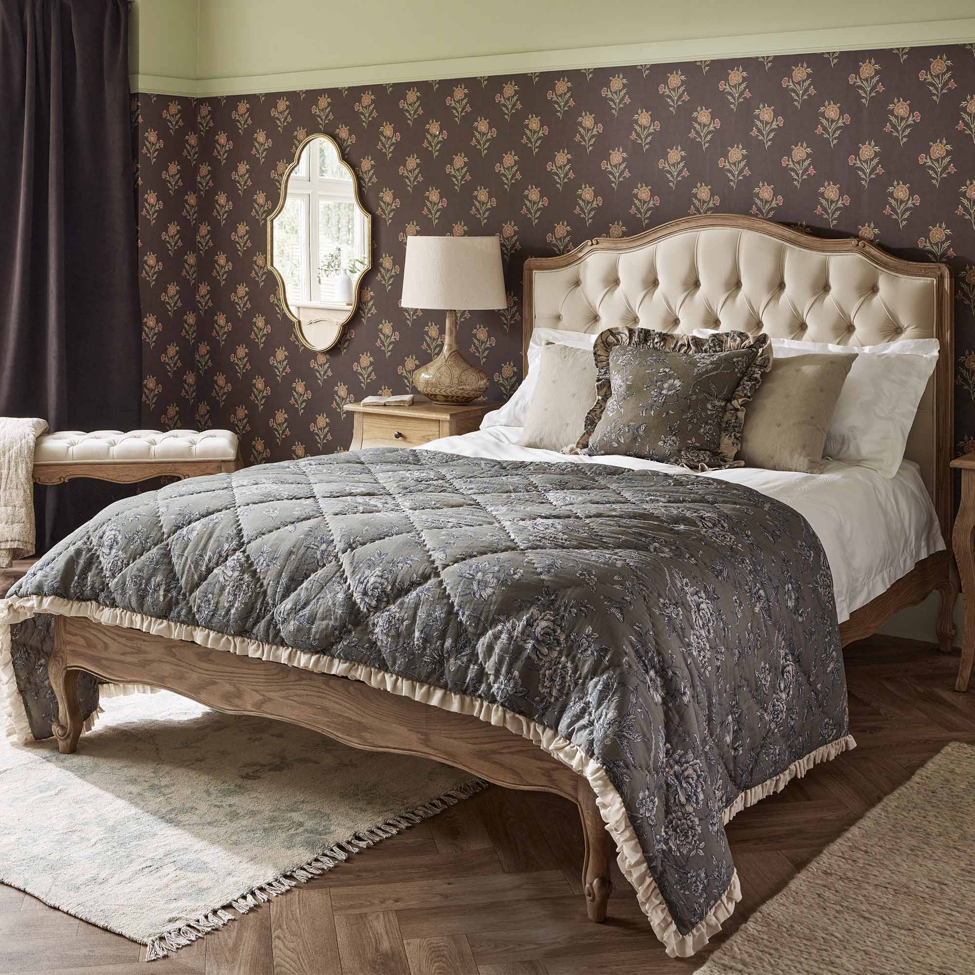 Elegant bedroom with a tufted headboard, floral wallpaper, and a quilted bedspread. A mirror, lamp, and curtains complement the decor.
