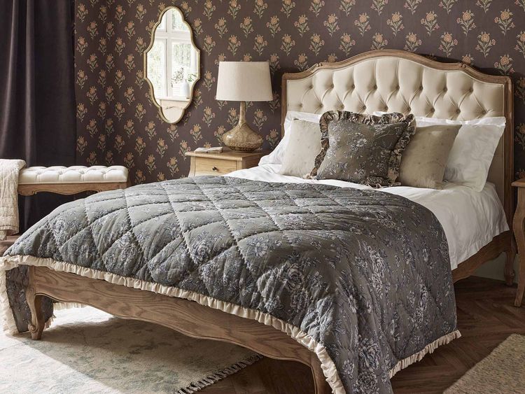 Elegant bedroom with a tufted headboard, floral wallpaper, and a quilted bedspread. A mirror, lamp, and curtains complement the decor.