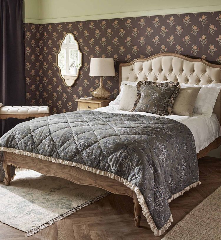Elegant bedroom with a tufted headboard, floral wallpaper, and a quilted bedspread. A mirror, lamp, and curtains complement the decor.