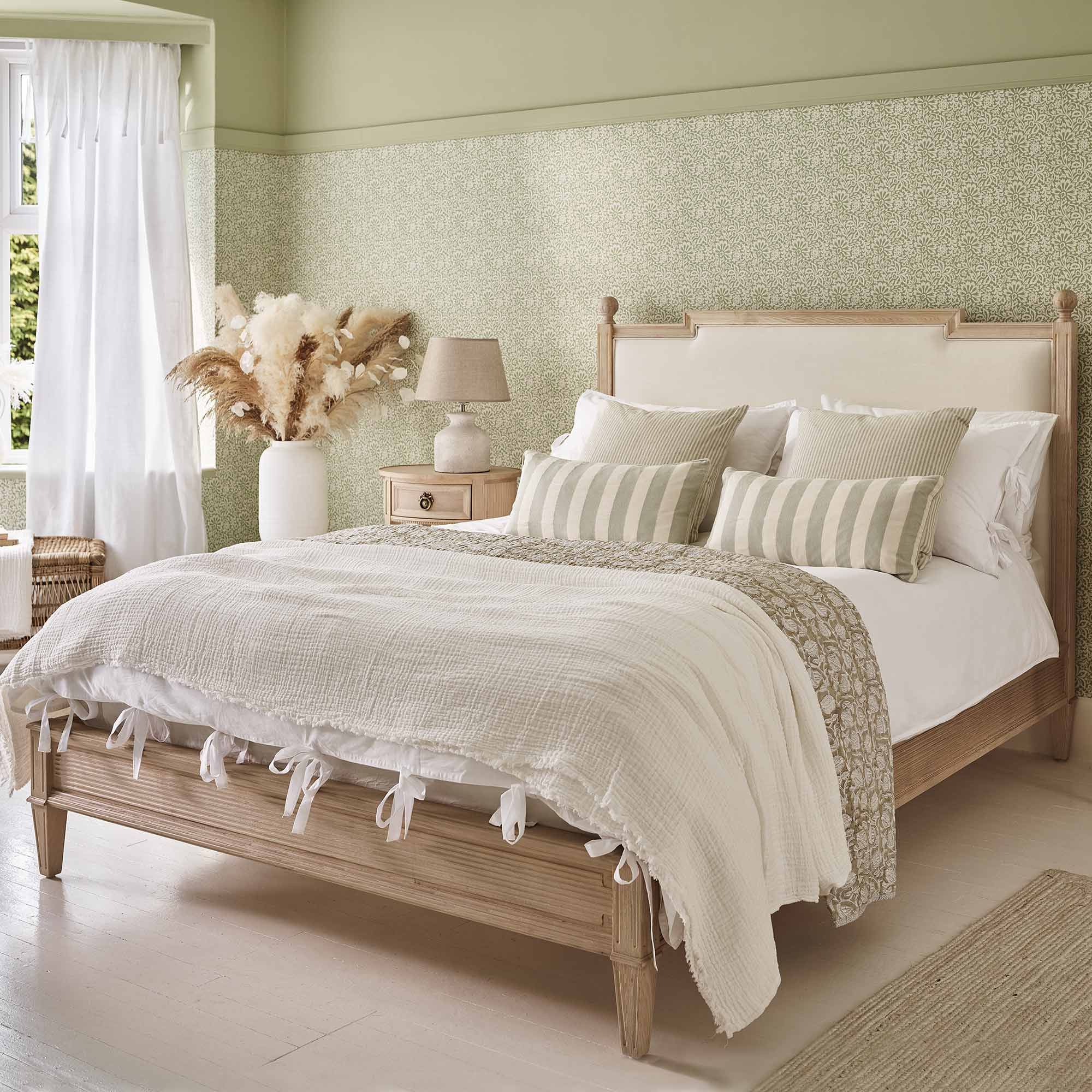 Cozy bedroom with a wooden bed, white and striped pillows, textured blankets, a bedside table, lamp, and dried flowers in a vase. Light green walls.