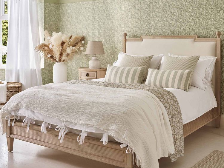 Cozy bedroom with a wooden bed, white and striped pillows, textured blankets, a bedside table, lamp, and dried flowers in a vase. Light green walls.