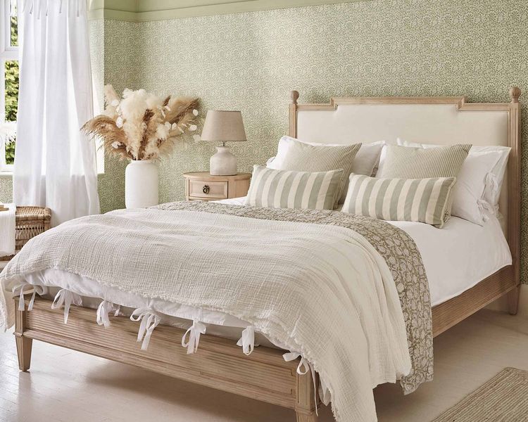 Cozy bedroom with a wooden bed, white and striped pillows, textured blankets, a bedside table, lamp, and dried flowers in a vase. Light green walls.