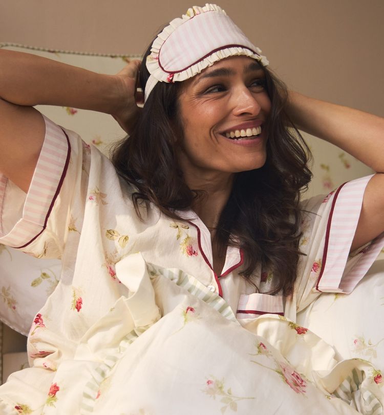 A woman smiling in bed, wearing floral pajamas and a striped sleep mask on her head, surrounded by matching floral-patterned bedding.