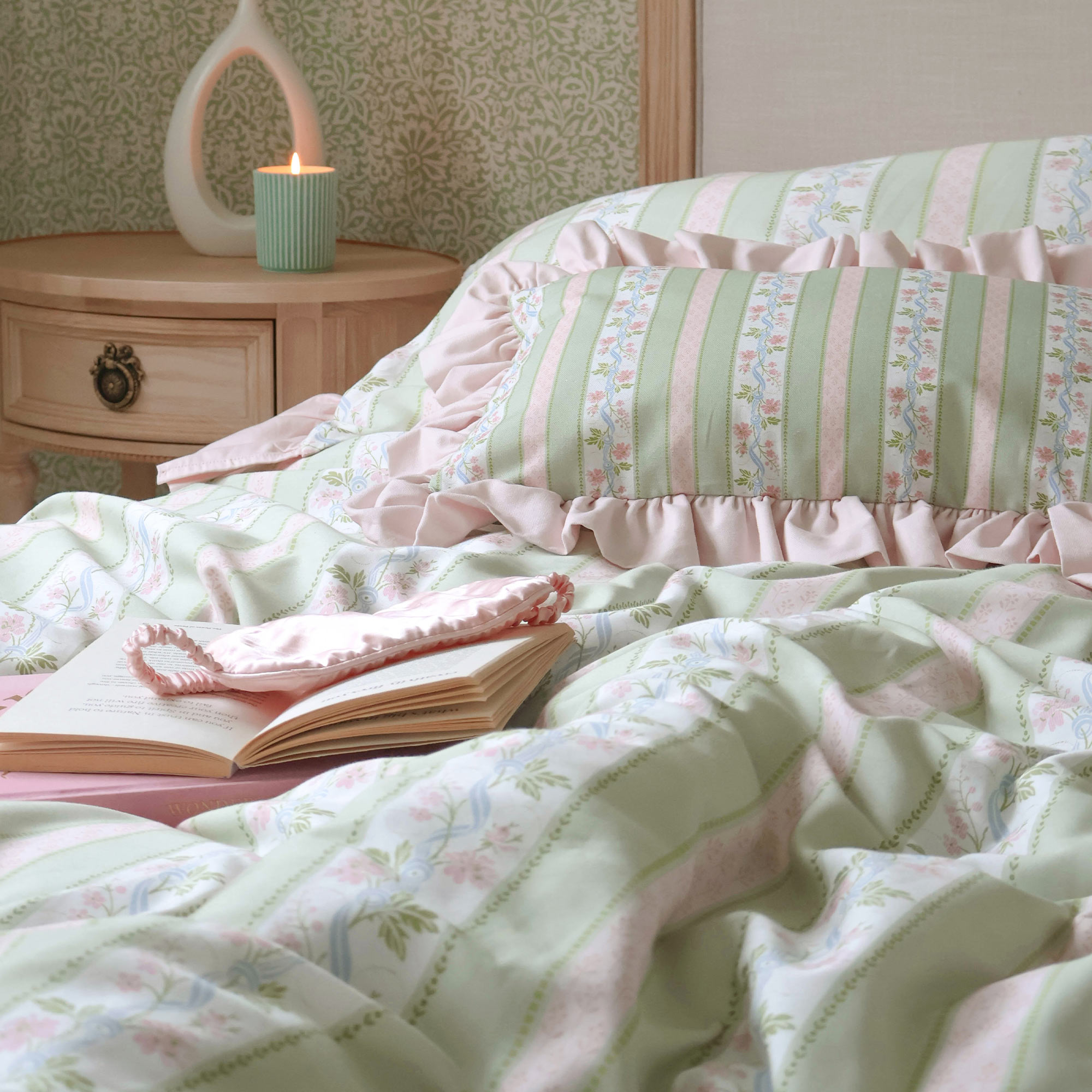 Cozy bedroom with pastel striped bedding, a ruffled pillow, an open book, and a lit candle on a wooden nightstand.