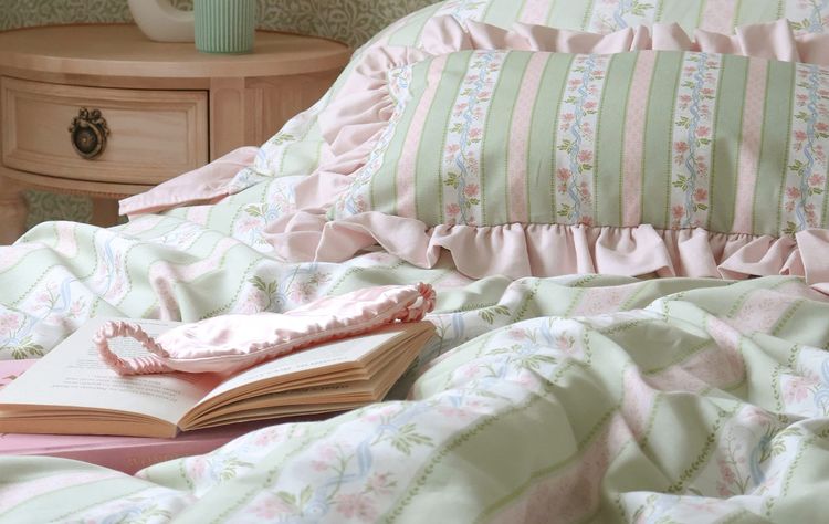Cozy bedroom with pastel striped bedding, a ruffled pillow, an open book, and a lit candle on a wooden nightstand.