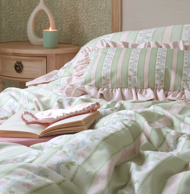 Cozy bedroom with pastel striped bedding, a ruffled pillow, an open book, and a lit candle on a wooden nightstand.