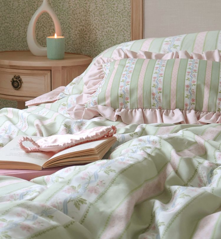 Cozy bedroom with pastel striped bedding, a ruffled pillow, an open book, and a lit candle on a wooden nightstand.