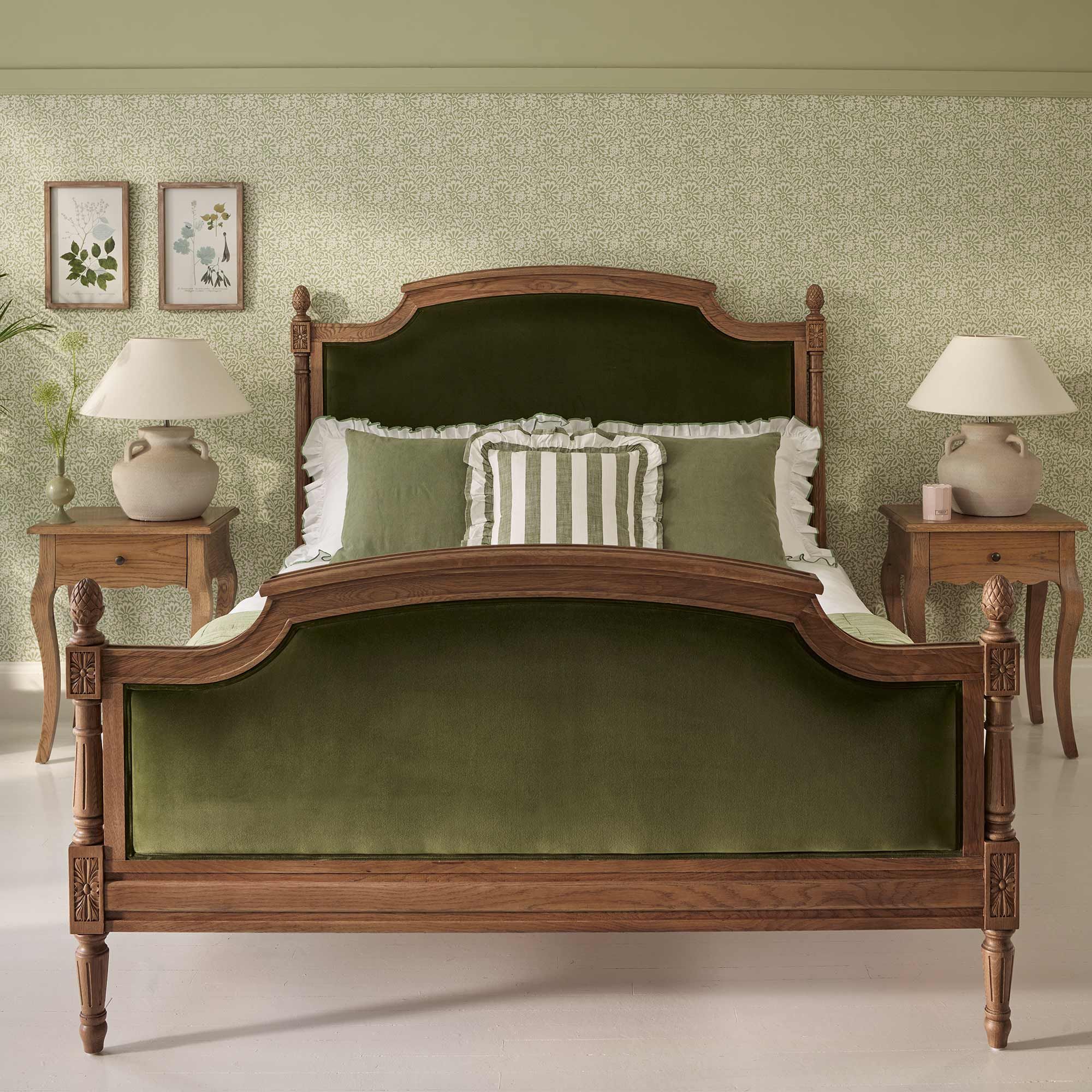 Elegant bedroom with a wooden bed frame and green velvet upholstery, flanked by two wooden nightstands with lamps, against patterned wallpaper.
