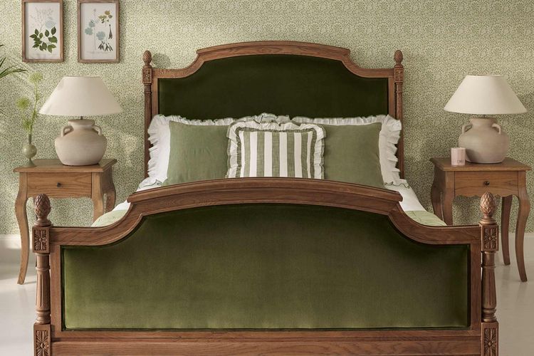 Elegant bedroom with a wooden bed frame and green velvet upholstery, flanked by two wooden nightstands with lamps, against patterned wallpaper.