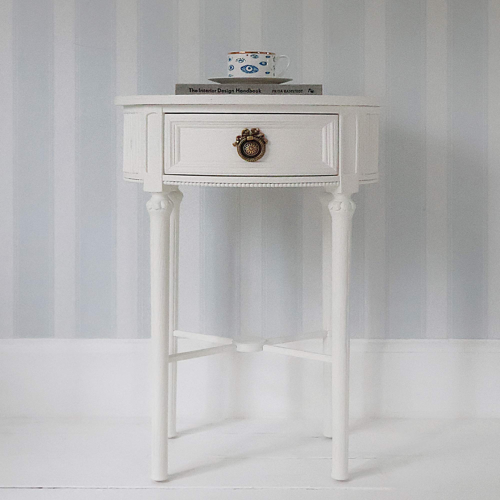 White round side table with a drawer and ornate handle, topped with a mug and a book, set against a light blue striped wall.
