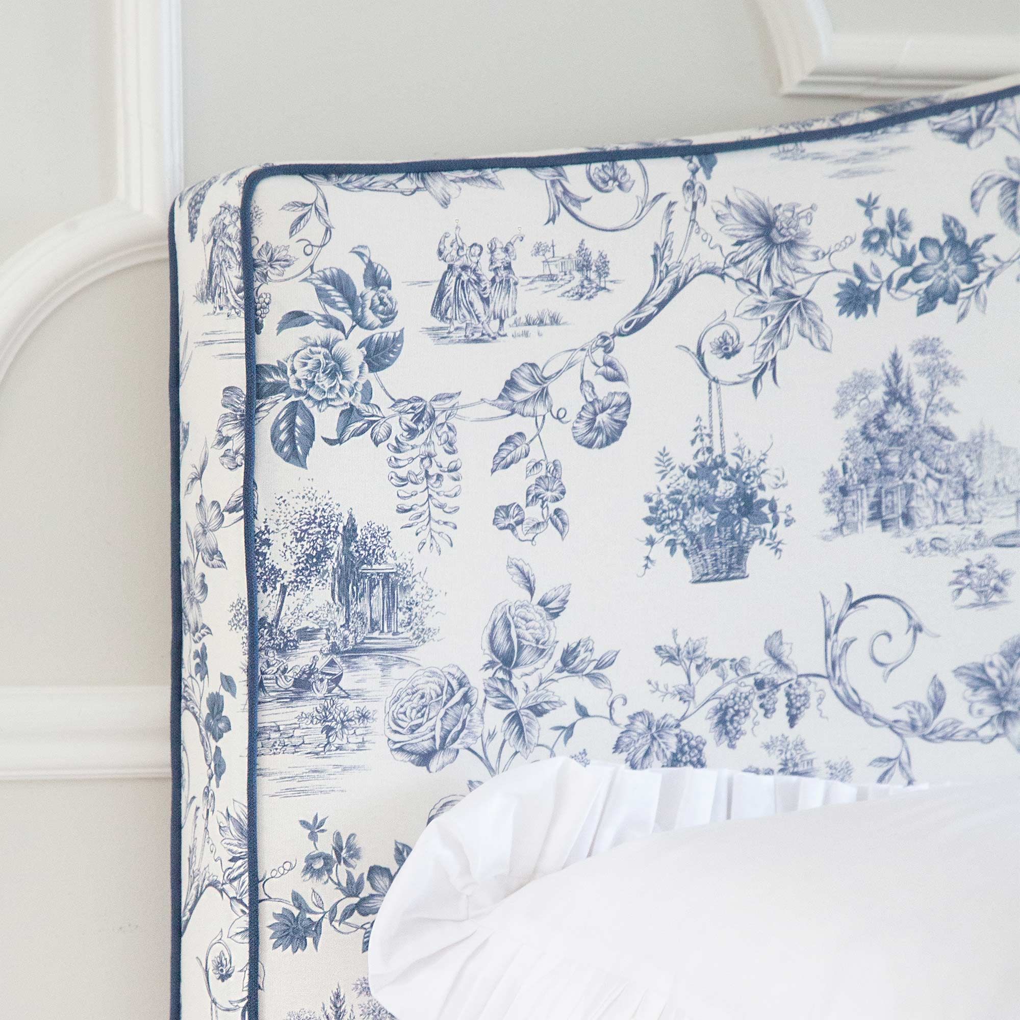 Close-up of a bed's corner with a blue and white floral and pastoral toile fabric headboard, and a white pillow with ruffled edges.