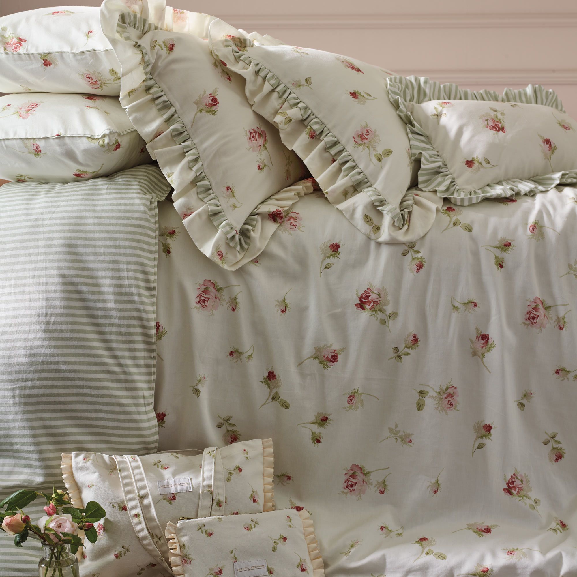 Floral and striped bedding with ruffled edges on a neatly made bed, featuring pink roses and green leaves.