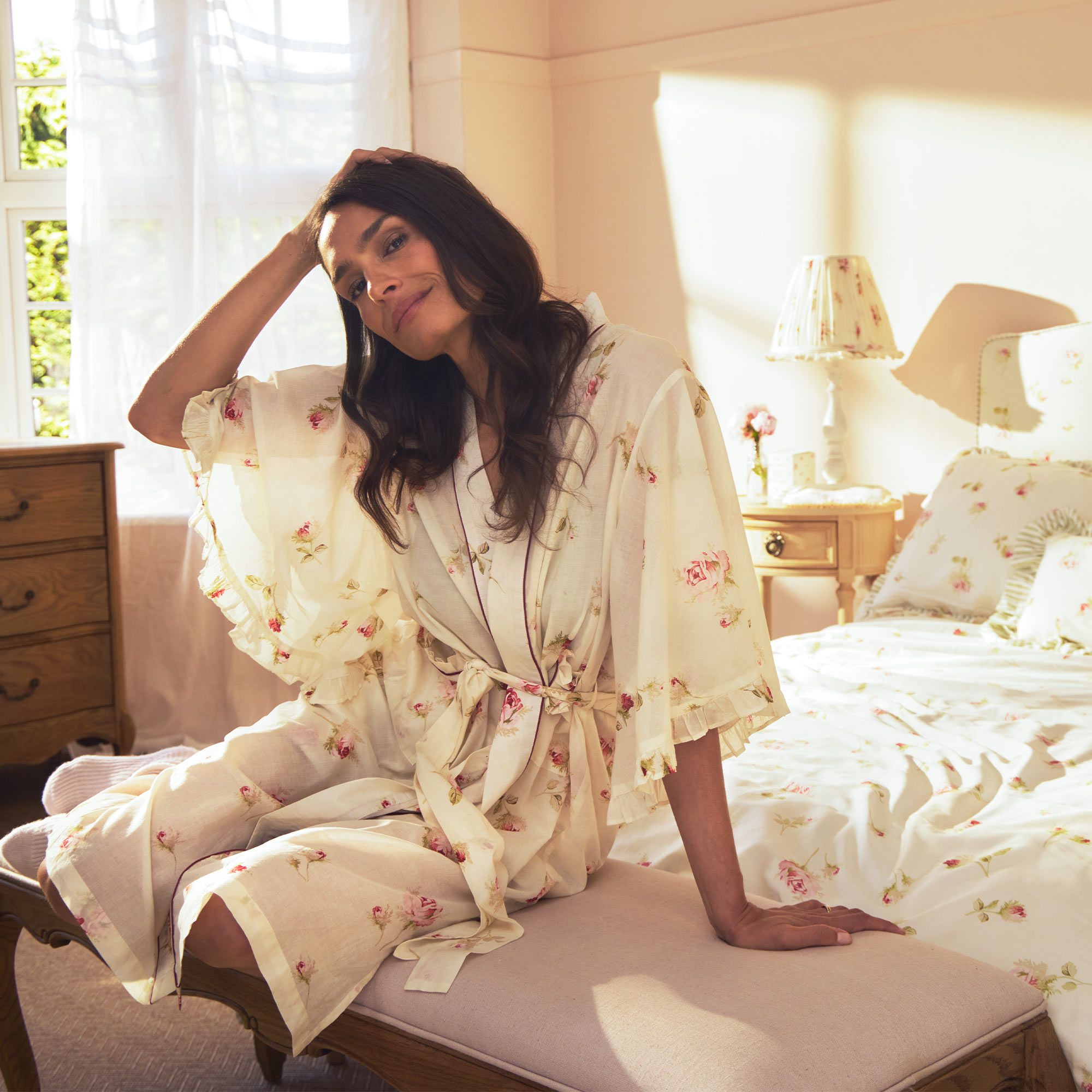 A woman in a floral robe sits on an upholstered bench in a sunlit bedroom with a floral bedspread and wooden furniture, smiling softly.