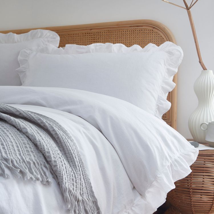 Cozy bed with white ruffled pillows and duvet, gray throw blanket, wicker headboard, and a white sculpted lamp on a wooden nightstand.