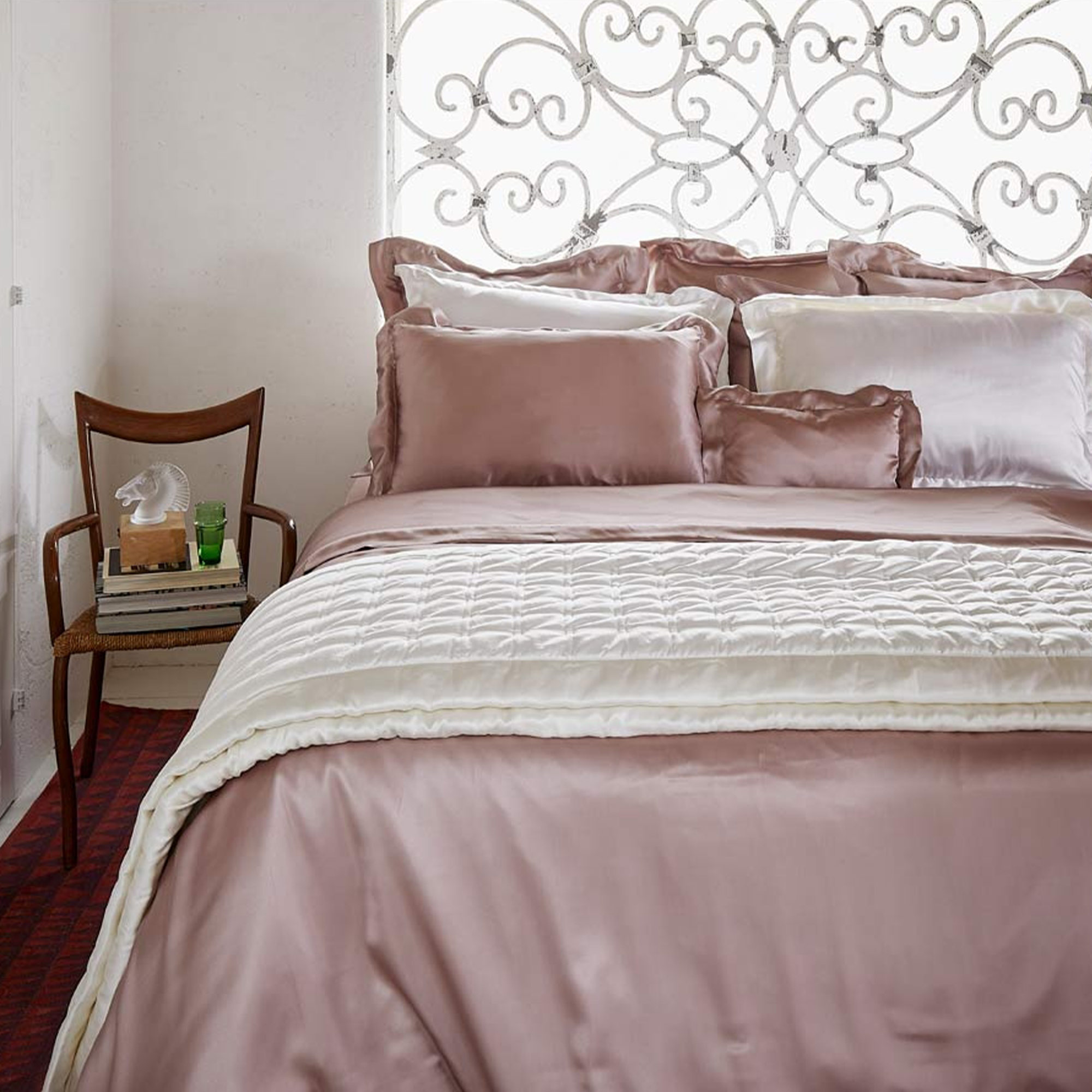Elegant bedroom with a neatly made bed featuring silky mauve and white bedding, decorative pillows, and a wooden chair with books and a glass.