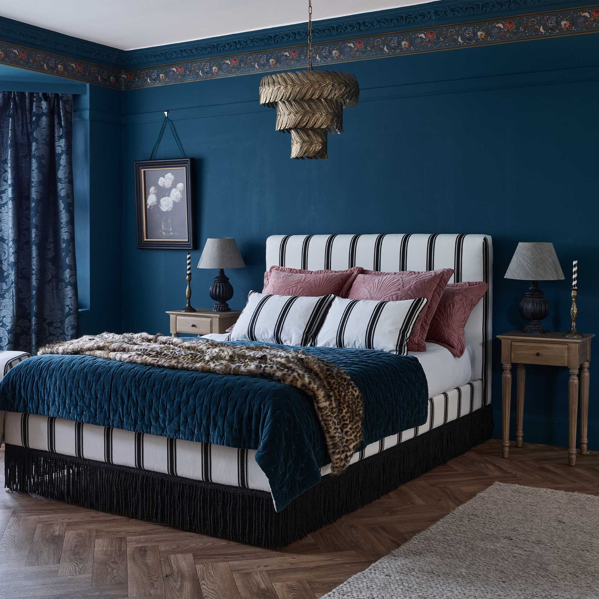 Elegant bedroom with a striped bed, blue and burgundy accents, wooden nightstands, patterned curtains, and a decorative chandelier.