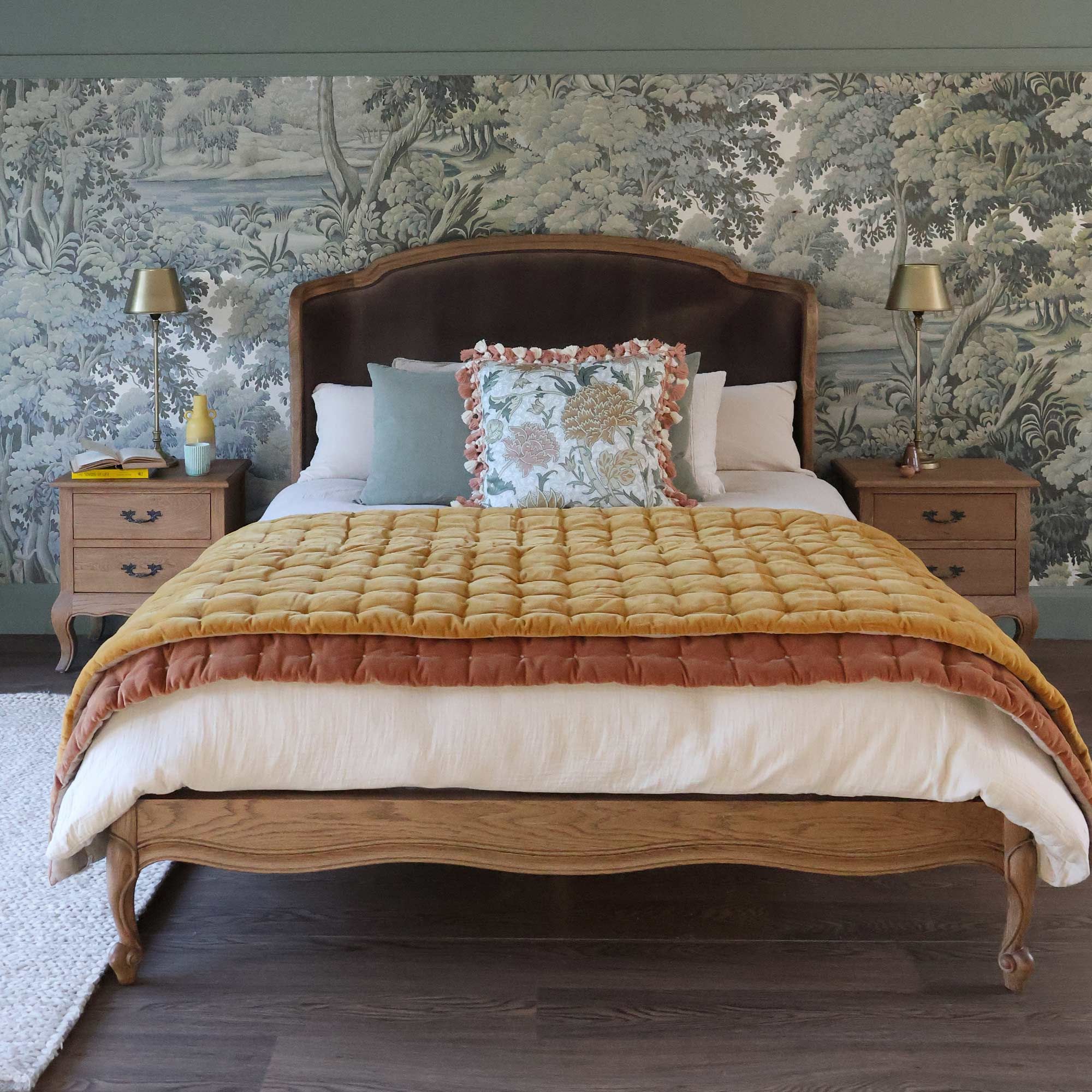 Elegant bedroom with a vintage bed, ornate headboard, yellow quilt, decorative pillows, wooden nightstands, and a nature-themed wallpaper.