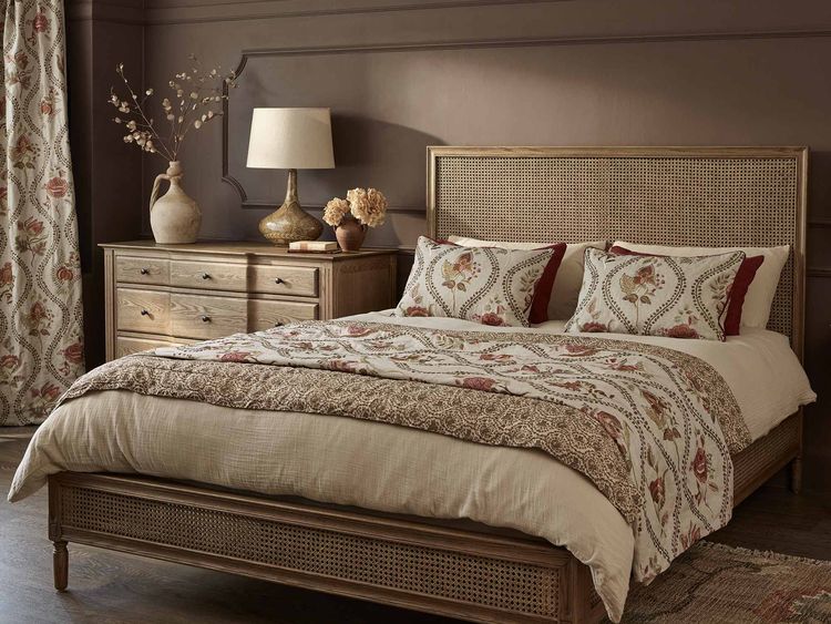 Cozy bedroom with a wooden bed, floral bedding, a matching dresser, lamp, and decorative items, set against a warm brown wall.