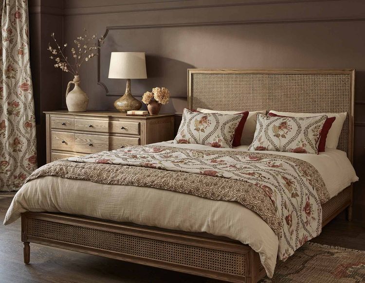 Cozy bedroom with a wooden bed, floral bedding, a matching dresser, lamp, and decorative items, set against a warm brown wall.