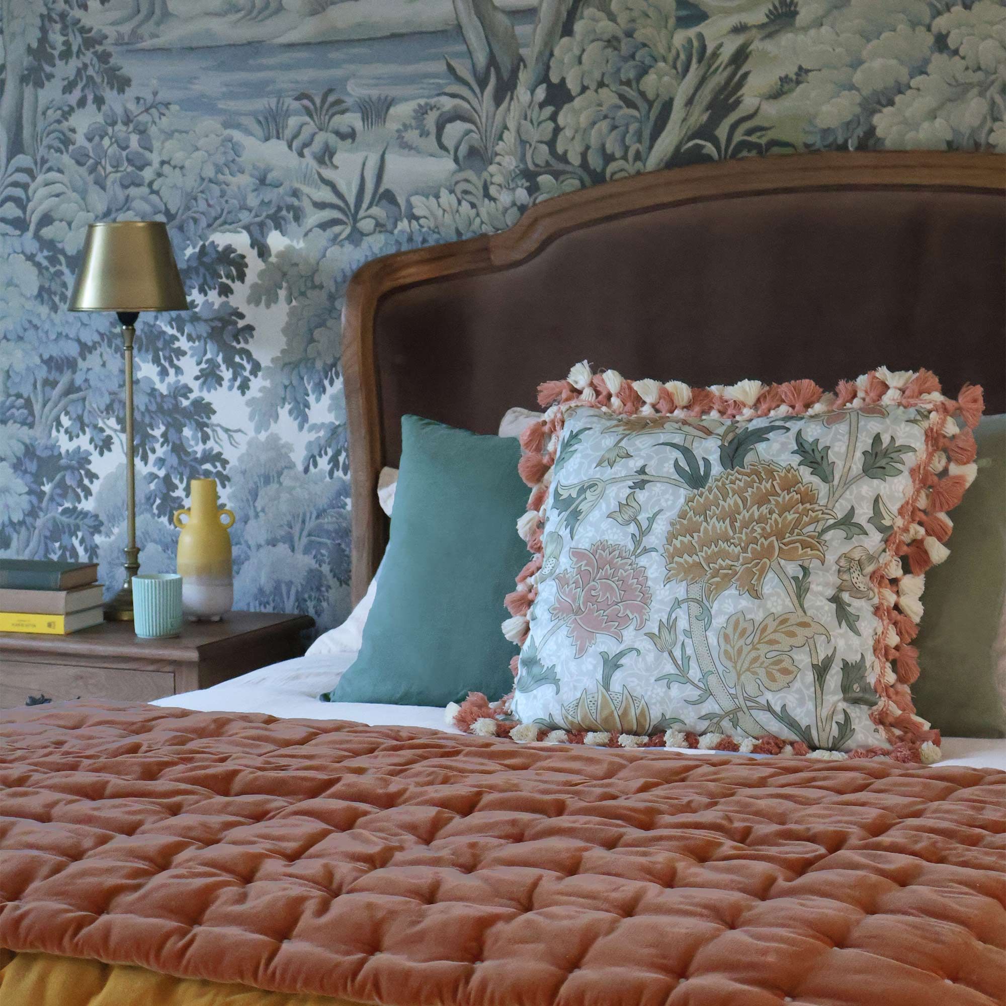 Cozy bedroom with a floral pillow, orange quilt, green cushions, and a wallpapered forest scene. A lamp and books are on the bedside table.