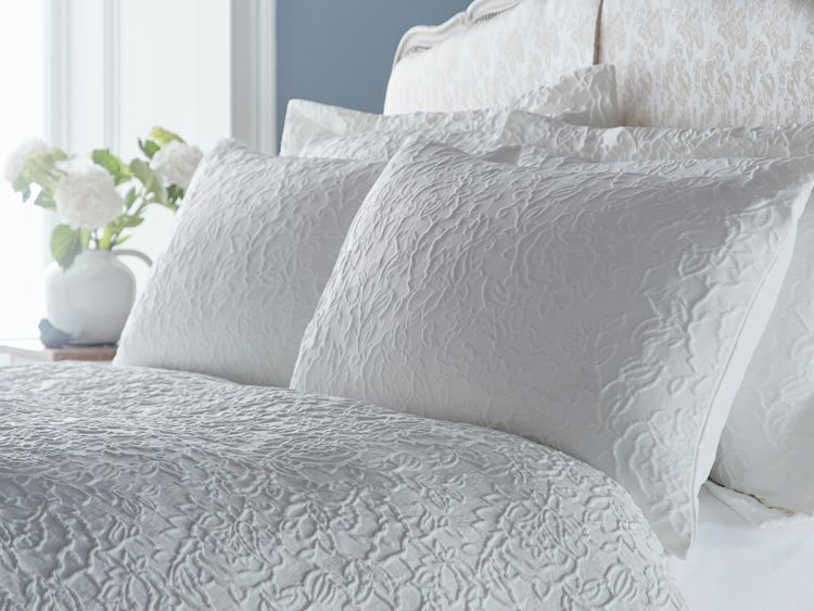 Elegant bedroom with a white, textured bedspread and pillows. A vase with white flowers is on the bedside table. Light blue wall in the background.