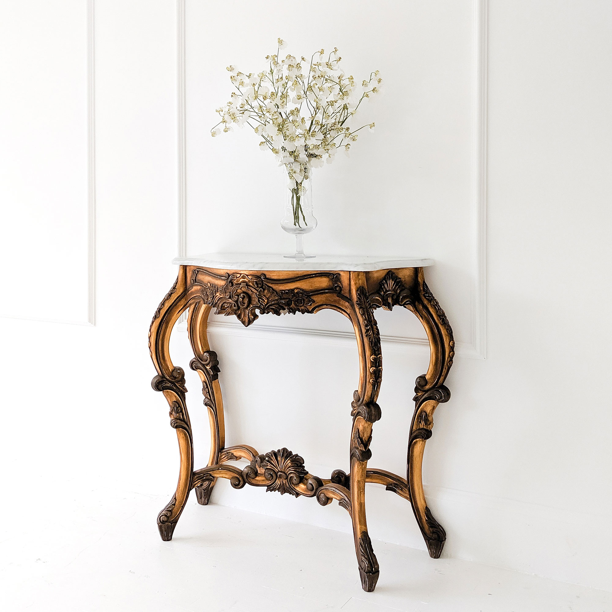 Ornate wooden console table with a white marble top, holding a glass vase of delicate white flowers, set against a white paneled wall.