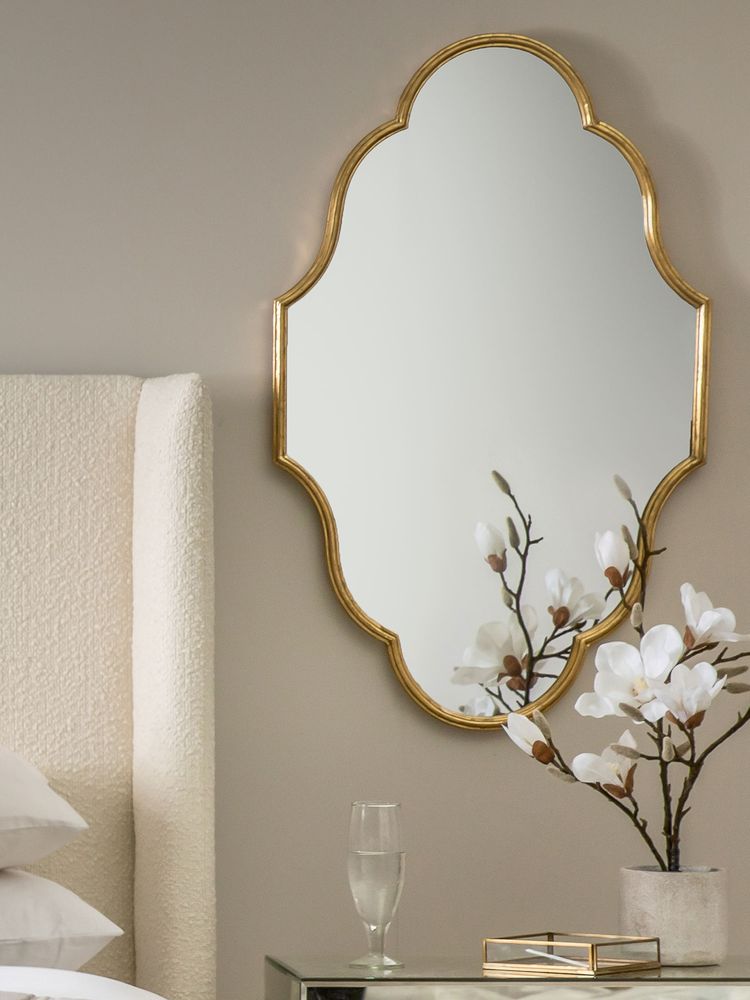 Elegant bedroom with a scalloped gold-framed mirror, magnolia flowers in a vase, and a glass of water on a mirrored nightstand.