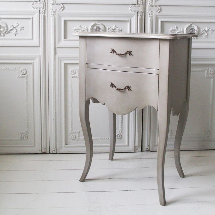 Elegant vintage-style gray nightstand with two drawers, ornate handles, and curved legs on a white wooden floor, against a decorative wall panel.