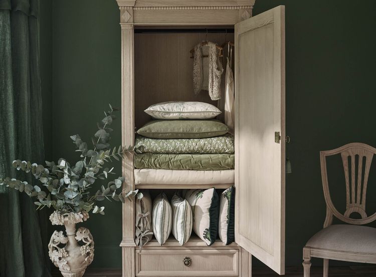 Open wooden wardrobe with neatly stacked green linens and pillows, next to a decorative vase with leaves and an elegant chair.