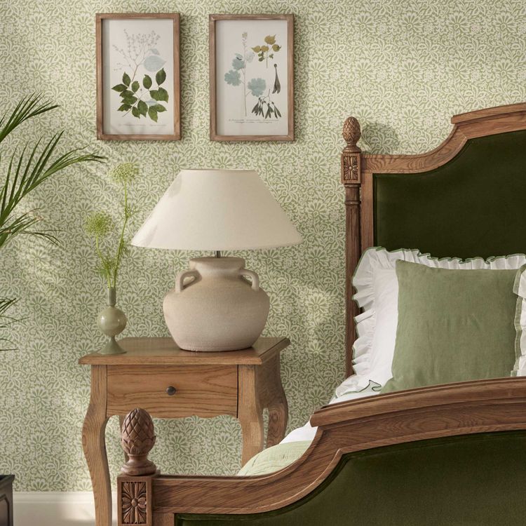 Elegant bedroom with a wooden bed, green velvet headboard, patterned wallpaper, bedside table with a lamp, and framed botanical art.