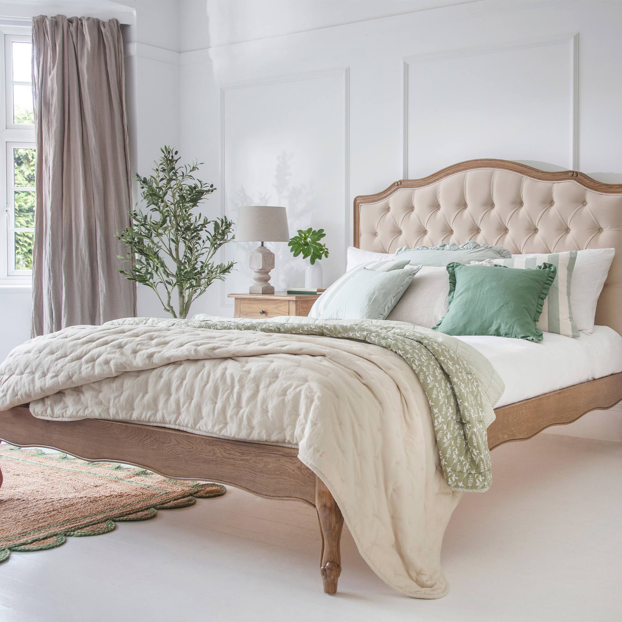 Elegant bedroom with a tufted headboard, white and green bedding, a wooden bed frame, and a potted plant by a window with beige curtains.