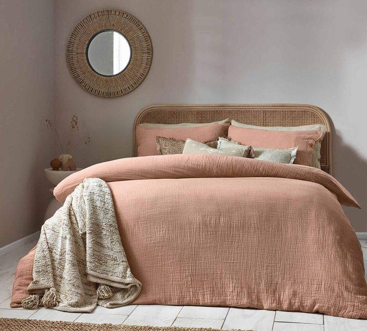 A cozy bedroom with a pink duvet, textured pillows, a woven headboard, and a round wicker mirror on a light pink wall.