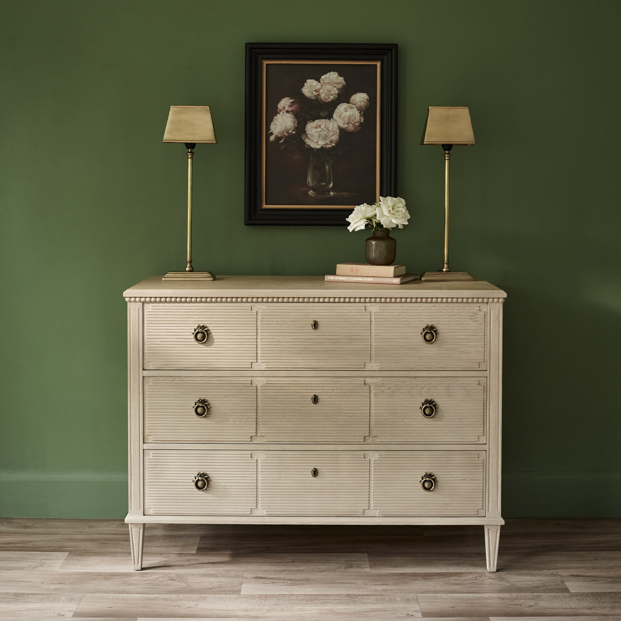 A beige dresser with six drawers, two lamps, and a flower painting on a green wall. A small vase with flowers sits on top.
