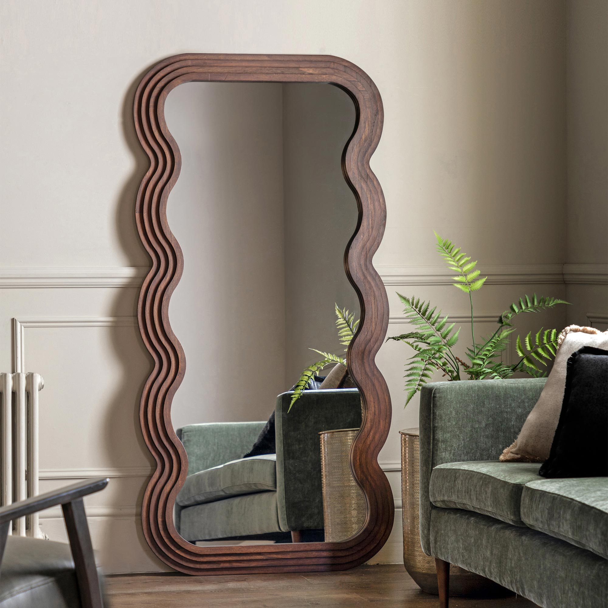 Wavy-edged wooden mirror leaning against a wall, reflecting a green sofa and plants in a stylish, minimalist room.