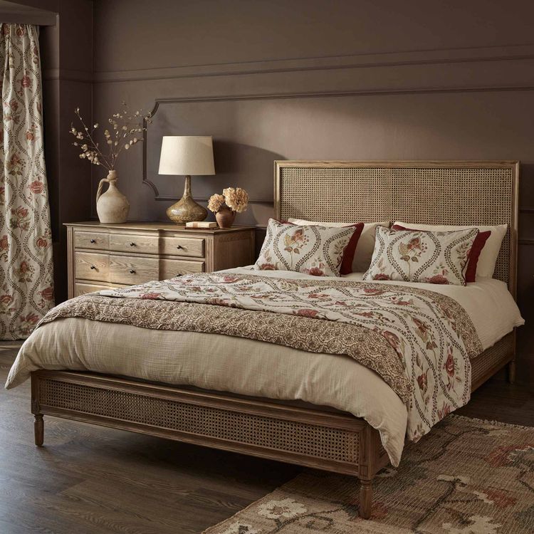 Cosy bedroom with a wooden bed, floral-patterned bedding, matching curtains, a dresser, a lamp, and decorative vases on a dark wood floor.