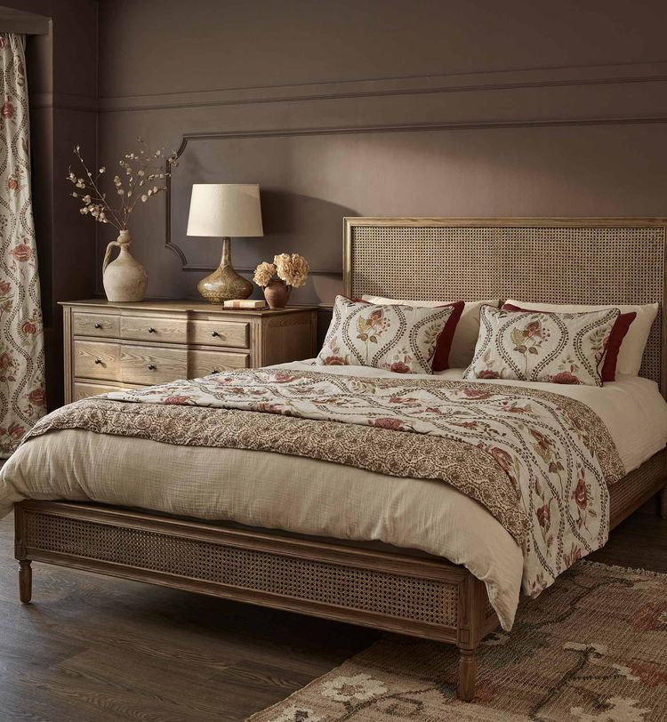 Cosy bedroom with a wooden bed, floral-patterned bedding, matching curtains, a dresser, a lamp, and decorative vases on a dark wood floor.