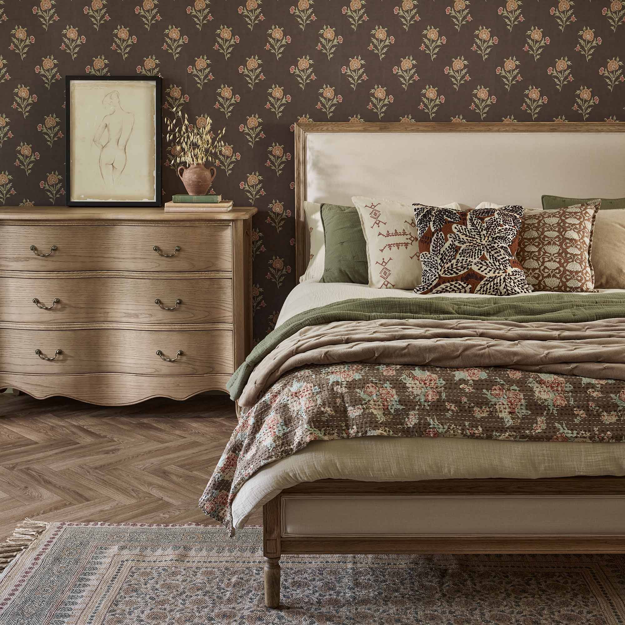 Cozy bedroom with a floral-patterned bed, wooden dresser, framed sketch, and floral wallpaper. Warm, earthy tones throughout the space.