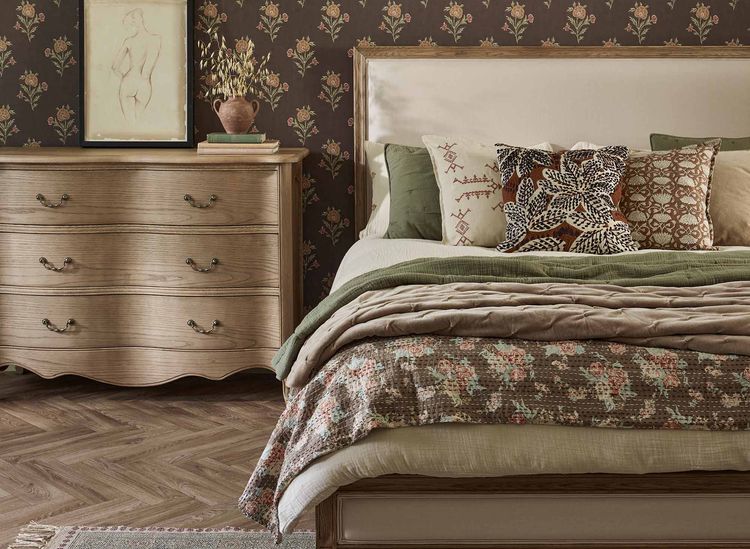Cozy bedroom with a floral-patterned bed, wooden dresser, framed sketch, and floral wallpaper. Warm, earthy tones throughout the space.