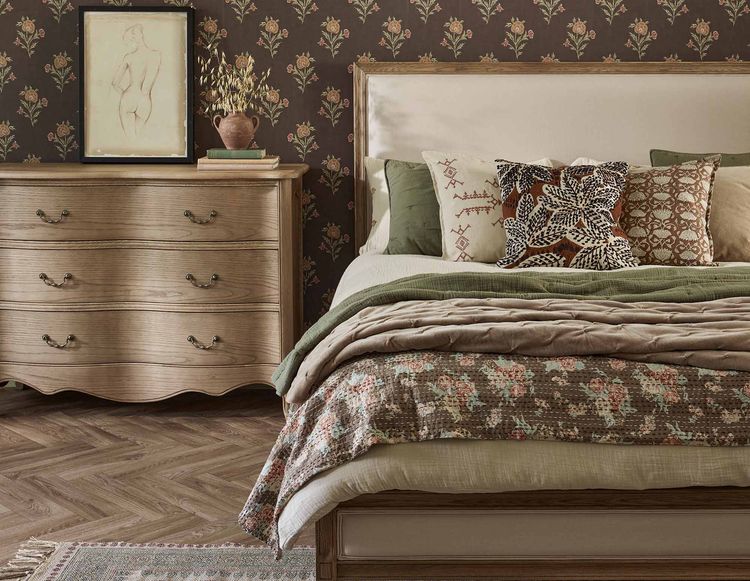 Cozy bedroom with a floral-patterned bed, wooden dresser, framed sketch, and floral wallpaper. Warm, earthy tones throughout the space.